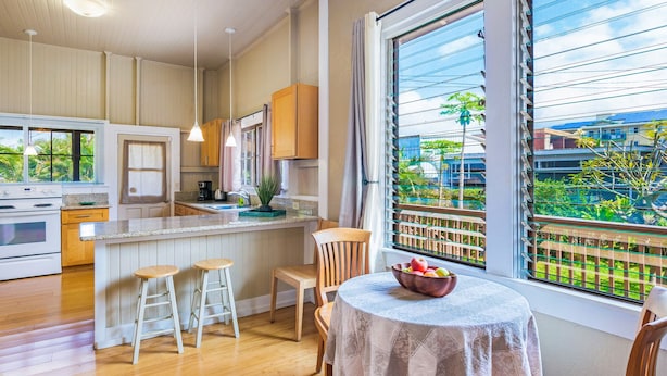 A modern kitchen with wooden cabinets, a central island with stools, a dining table with a tablecloth, and large windows with a view of greenery.