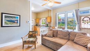 A living room with a sofa, a chair, a ceiling fan, and a kitchen in the background.