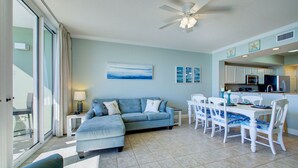 A living room with a blue sofa, dining table, and chairs, a ceiling fan, and a kitchen area with cabinets and appliances.