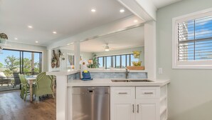 Una cocina moderna con armarios blancos, lavavajillas de acero inoxidable y un amplio ventanal con persianas.