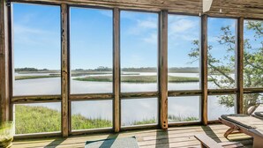 Habitación con amplias ventanas que dan a una masa de agua tranquila y humedal. Se observan bancos de madera y un ventilador de techo.