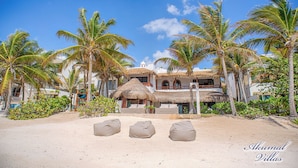 Un complejo turístico en la playa con edificios de techo de paja, palmeras y tres sillones de arena.