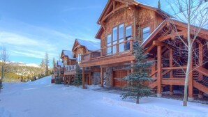 A large wooden lodge with multiple balconies and a stone foundation, surrounded by snow and trees.