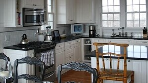 A kitchen with white cabinets, a microwave, a stove, and a dining area with chairs and a table. - The Sandbox   Beautiful Home in Tom Nevers (Nantucket)