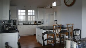 A kitchen with white cabinets, a black countertop, a wooden dining table with chairs, and a clock on the wall. - The Sandbox   Beautiful Home in Tom Nevers (Nantucket)