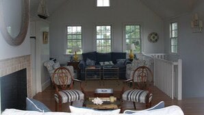 A spacious living room with a high ceiling, a fireplace, a large mirror, and a view of the outdoors through multiple windows. - The Sandbox   Beautiful Home in Tom Nevers (Nantucket)