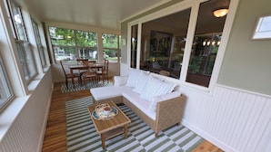 Eine überdachte Veranda mit einem Korbsessel, einem Esstisch mit Stühlen und Blick ins Freie.