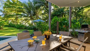 An outdoor dining area with a table set for two, surrounded by lush greenery and a pool.