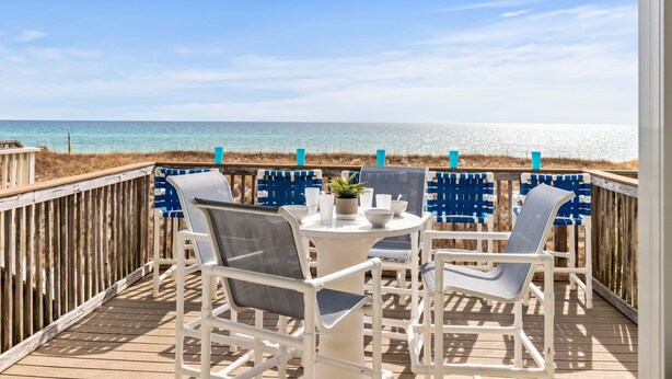 Eine Holzterrasse mit weißen Stühlen und einem Tisch, mit Blick auf einen Strand.
