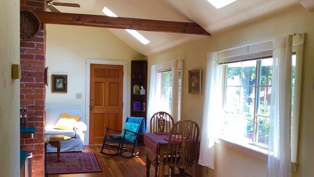 A spacious living area with a wooden floor, a dining table with chairs, a rocking chair, a bench, and a large window with curtains. - Garden Cottage on College Hill (Eugene)
