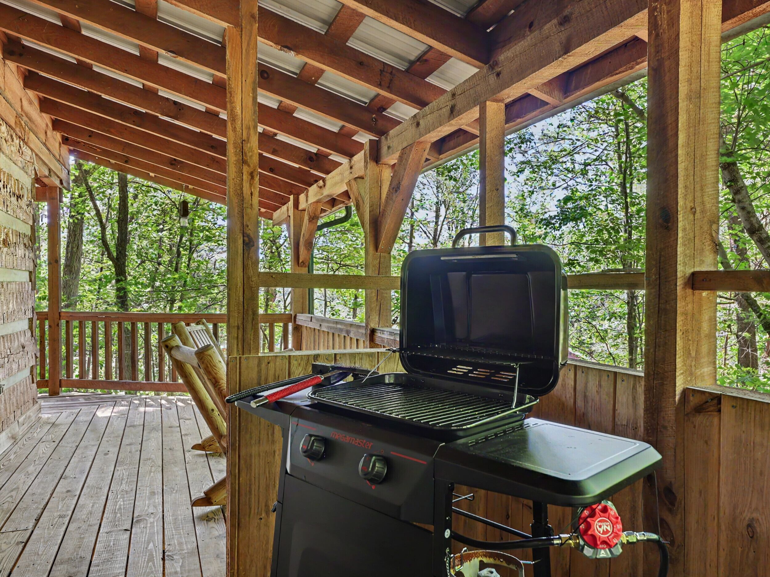 Ein überdachter Außenbereich mit tragbarem Gasgrill, Holzdeck und Blick auf Bäume.