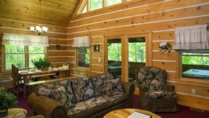 Ein Blockhaus-Interieur mit Holzbalkendecke, einer Couch mit Waldmuster, einem Esstisch mit Stühlen und einem Kamin.