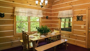 Eine Holzhütte mit Esstisch, gedeckter für vier Personen, zwei Fenster mit Vorhängen, einer Chandelieber und einer Bär-Dekoration an der Wand.