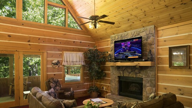 Ein gemütliches Blockhaus-Interieur mit Kamin aus Stein, Holzwänden, einem darüber montierten Fernseher und einer komfortablen Sitzecke mit Sofa und Couchtisch.