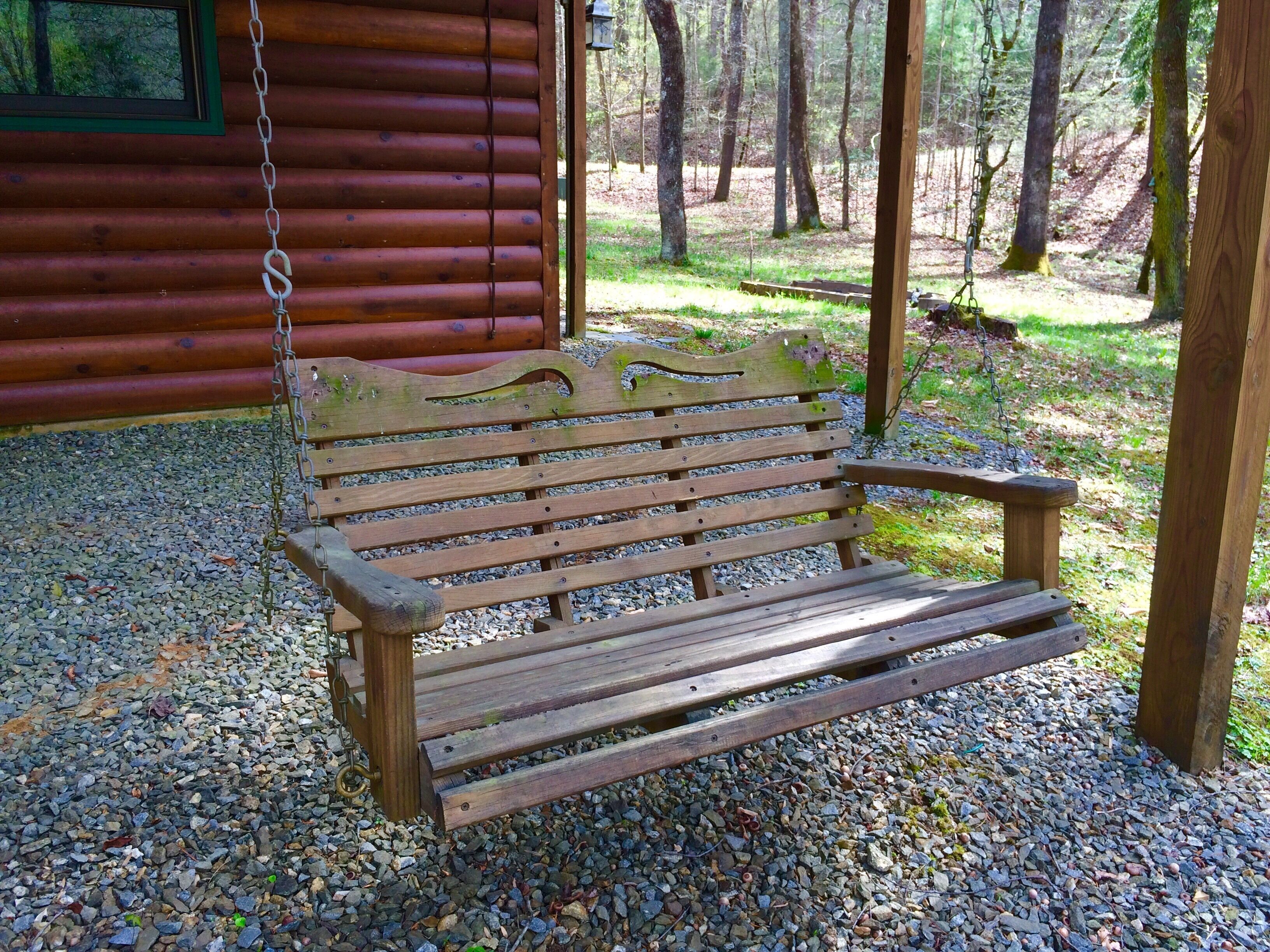 A wooden bench with a metal chain, set on a gravel path in front of a wooden cabin with a window.