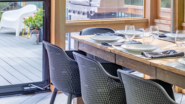 Un área de comedor al aire libre con mesa y sillas de madera, ventanas amplias y vistas a una parrilla y una terraza.