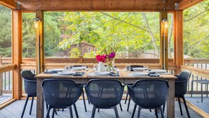 Una terraza de madera con una mesa preparada para cuatro personas, rodeada de sillas y vistas a árboles y una casa al fondo.