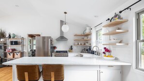 Una cocina moderna con armarios blancos, una isla central con dos taburetes altos, piso de madera y un amplio ventanal que proporciona luz natural.