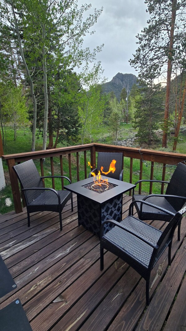 A deck with a fire pit table and four chairs, surrounded by trees and a mountain in the background.