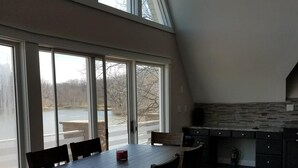 Un espace repas avec une grande table et des chaises, une cuisine dotée de meubles sombres et de grandes fenêtres offrant une vue sur un plan d’eau et des arbres.