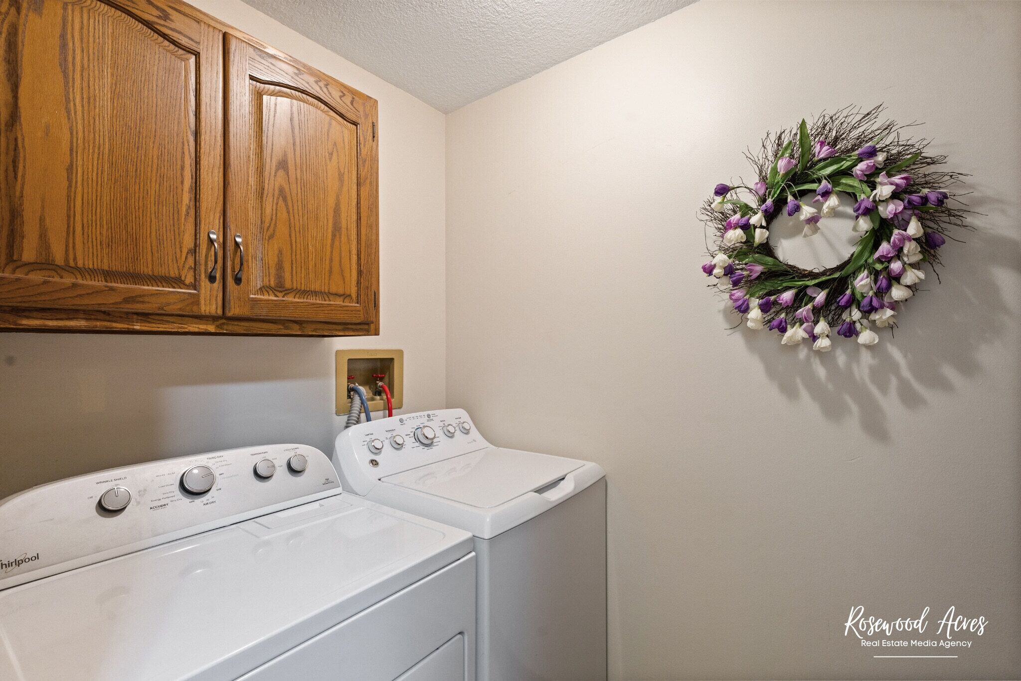 Een wasruimte met een witte wasmachine en droger, houten kastjes en een decoratieve krans aan de muur.