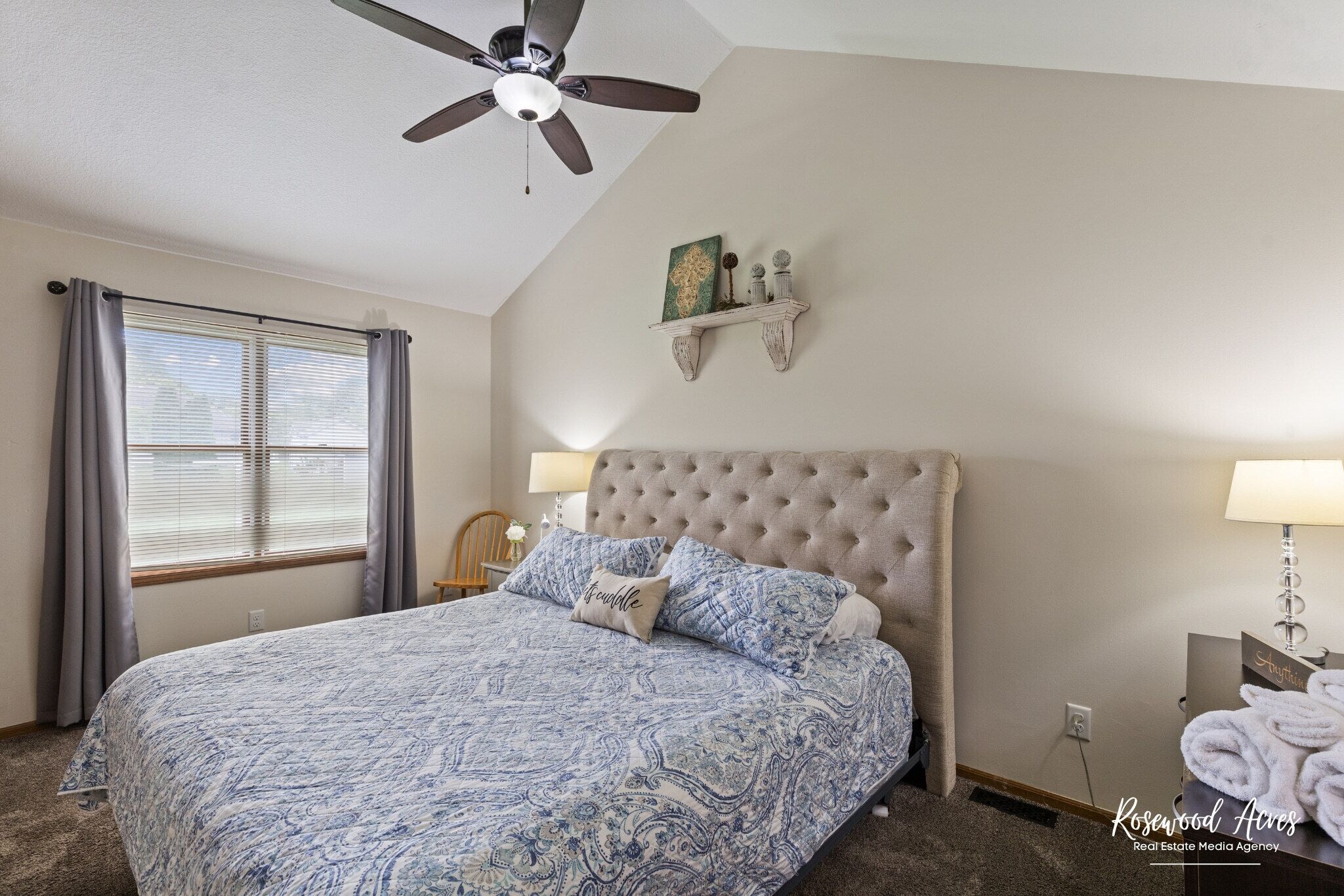 A bedroom with a tufted headboard, a bed with a blue and white patterned bedspread, a ceiling fan, a window with curtains, and a nightstand with a lamp and towels.