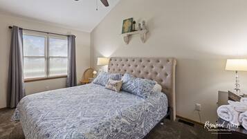 A bedroom with a tufted headboard, a bed with a blue and white patterned bedspread, a ceiling fan, a window with curtains, and a nightstand with a lamp and towels.