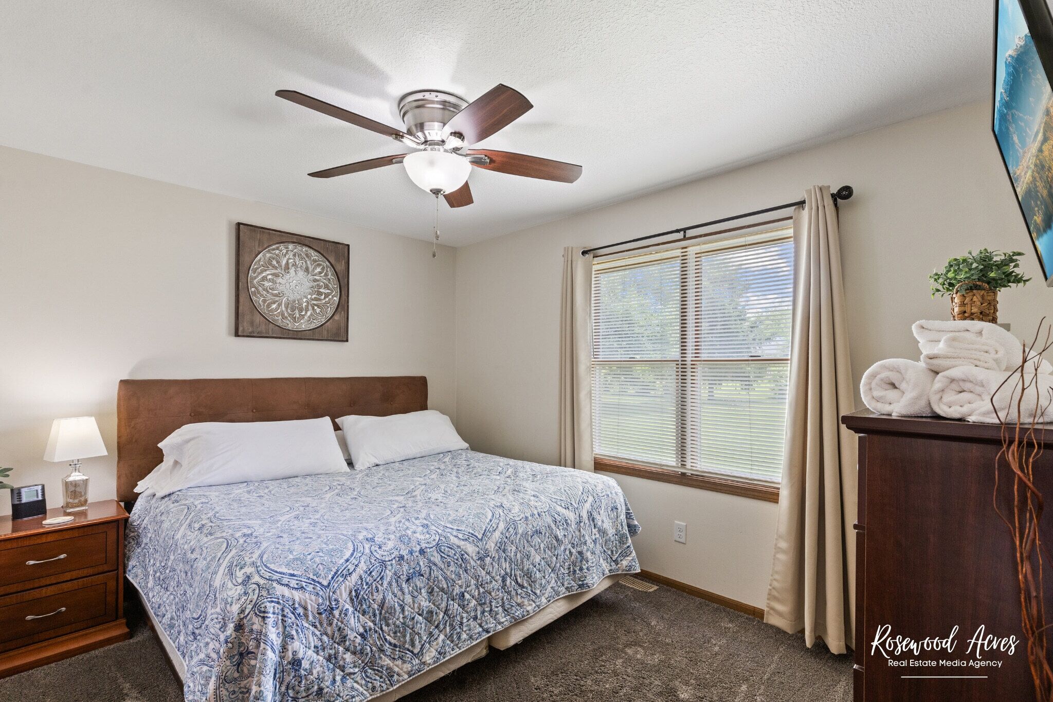 A bedroom with a bed, a ceiling fan, a window with curtains, a nightstand with a lamp, and a painting on the wall.