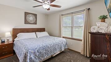 A bedroom with a bed, a ceiling fan, a window with curtains, a nightstand with a lamp, and a painting on the wall.