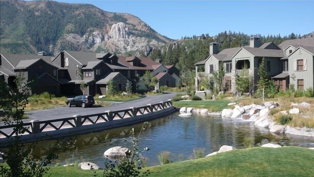 A residential area with houses, a bridge over a pond, and mountains in the background.