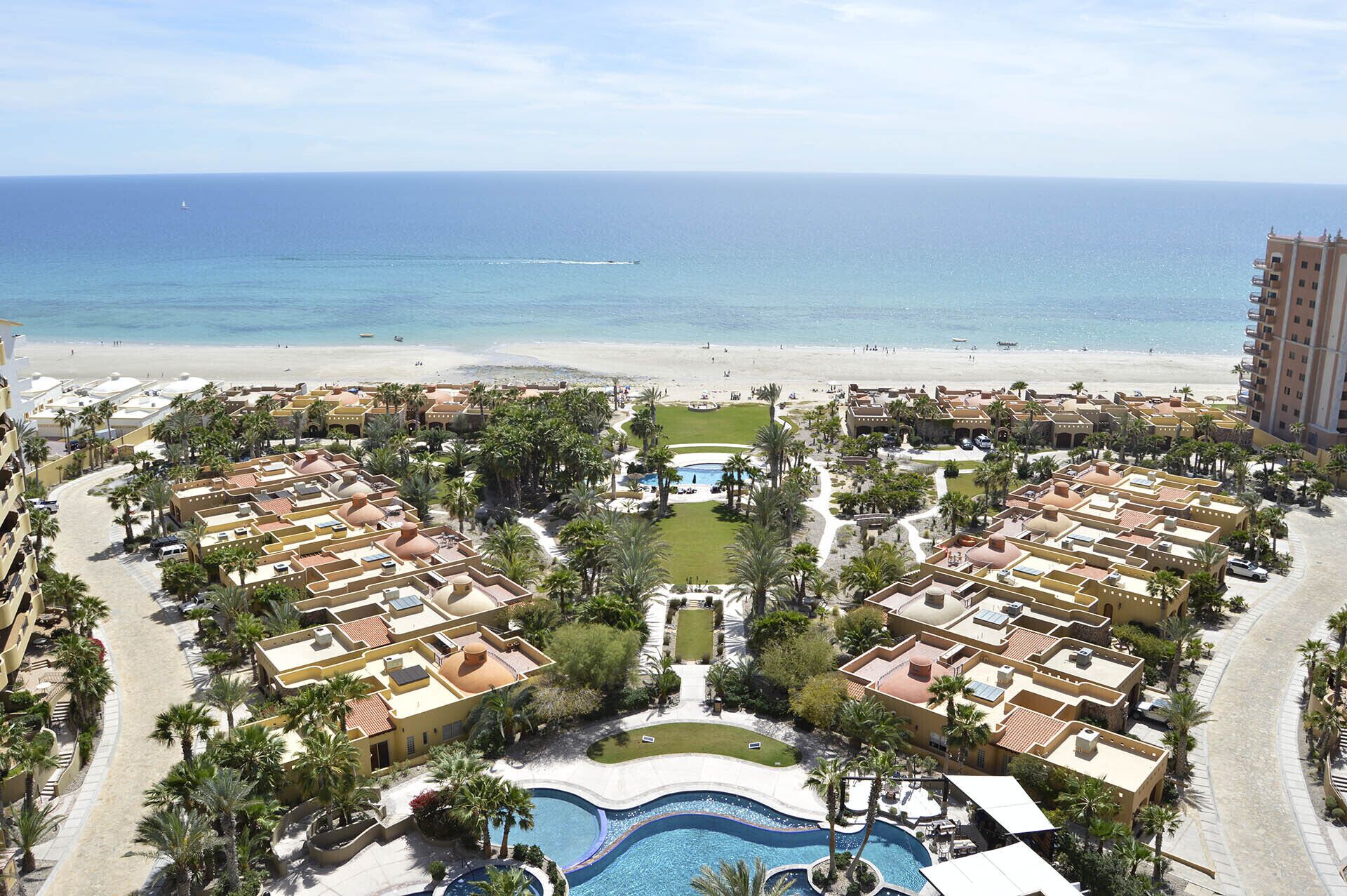 A resort with multiple buildings, a central pool area, and a beach in the background.