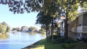 Vista su un lago con una zona erbosa, alberi e un edificio con un balcone.