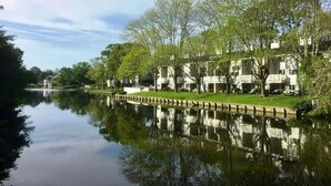 Un lago calmo con il riflesso di alberi e edifici, immerso nel verde e sotto un cielo sereno.