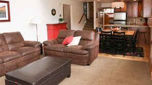 A living room with two brown leather sofas, a coffee table, and a red cabinet. There is a kitchen area with a dining table and chairs, and a staircase leading to another level. - 4 BR/4 BA Townhome - Spacious, comfortable, the perfect getaway! (Park City)