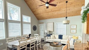 A spacious living area with a high ceiling, wooden beams, a dining table with chairs, a sofa, and a kitchen area with a countertop. - Lakeside Retreat- Family and Pet Friendly  (Fort Ann)