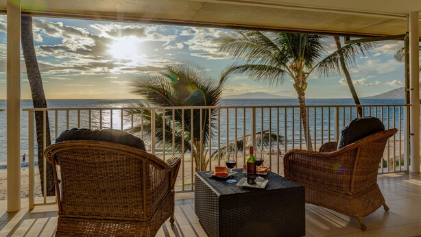 A balcony with wicker furniture, a table with a bottle and glass, and a view of the ocean and palm trees.