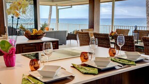 A dining table set with a bowl of fruit, wine glasses, and cutlery, overlooking the sea.