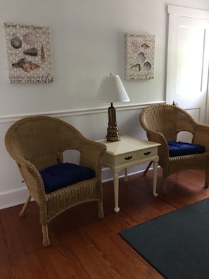 Two wicker chairs with blue cushions, a small white table with a lamp, and decorative wall art.