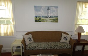 A living room with a brown sofa, two pillows with a ship wheel design, a framed painting of a lighthouse, a small table with a tray, and a lamp on a side table.