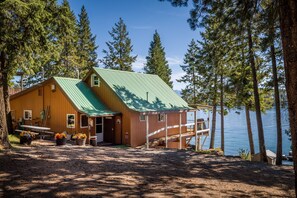 Une cabane dotée d’un toit végétalisé, de murs en bois et d’une jetée s’avançant dans un lac, le tout entouré d’arbres.