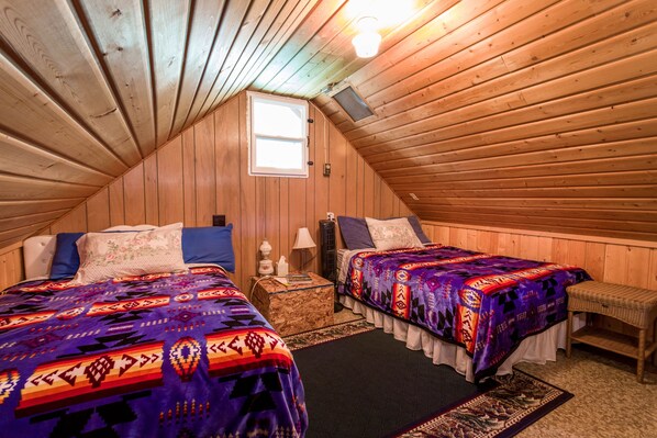 Een gezellig slaapkamer op zolder met houten wanden, twee bedden met kleurrijke beddengoed, een klein raam en een nachtkastje met een lamp.