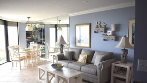 A living room with a sofa, coffee table, and dining area with chairs. There is a mirror, decorative items on the wall, and a lamp on a side table. - 3BR/2BA Oceanfront- Gorgeous Southern View-Gorgeous Kitchen & Baths-Book Early! (Myrtle Beach)