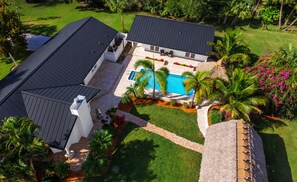Una propiedad residencial con piscina, patio cubierto y jardín bien cuidado con palmeras y plantas con flores.
