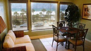 A room with a sofa, dining table, chairs, a large plant, and a view of the beach and ocean through large windows. - Stunning Sunset Views-Steps to Siesta Key Beach-A Couples' Romantic Beach Condo (Siesta Key)