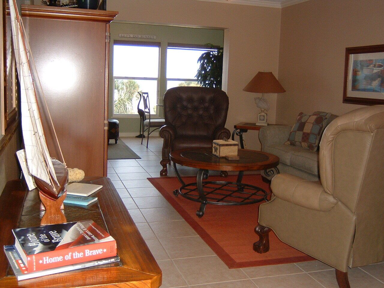 A living room with a brown leather armchair, a beige sofa, a wooden coffee table, and a red rug. There is a bookshelf with books and a small table with a lamp.