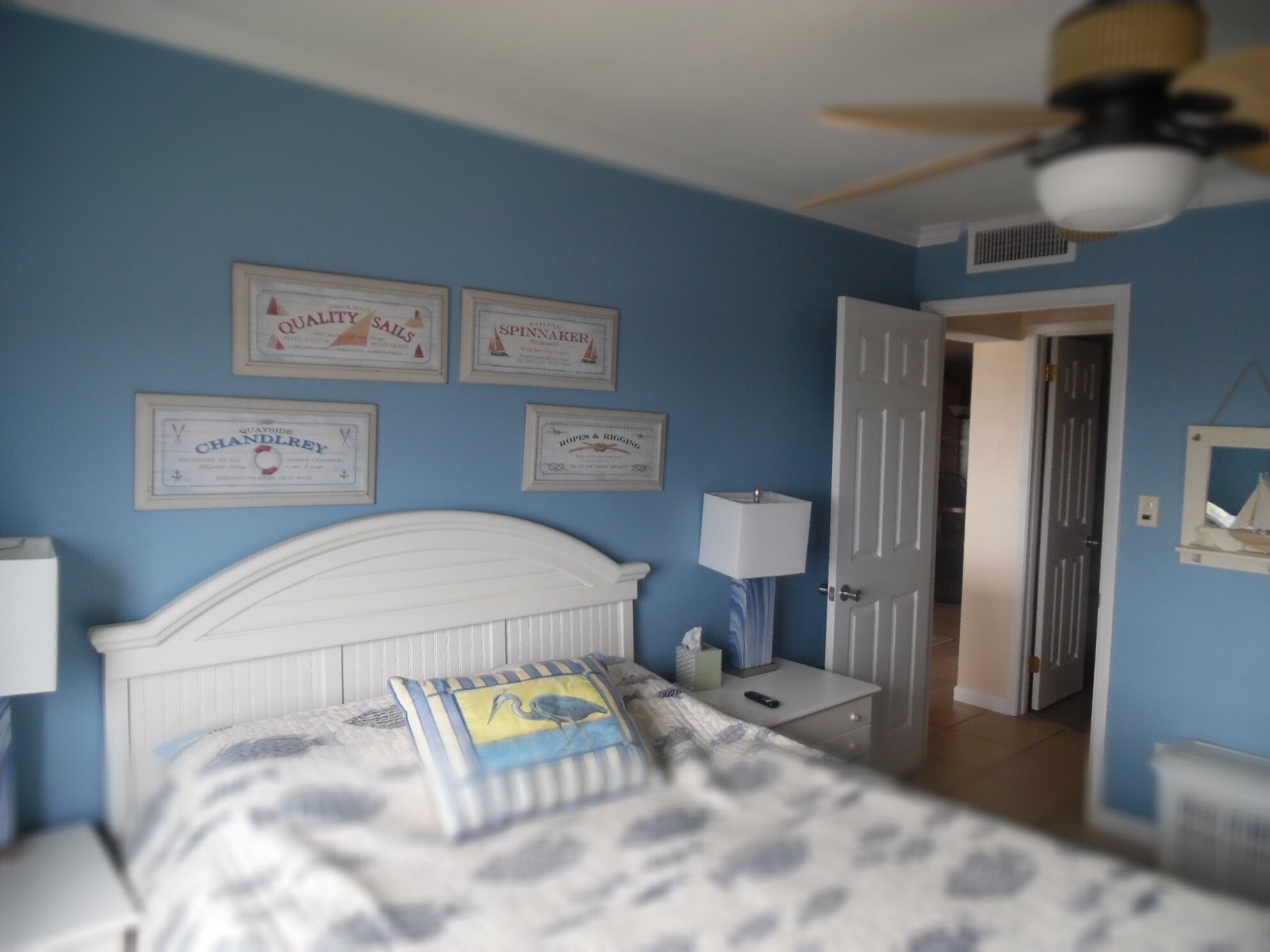 Un dormitorio con cabecera blanca, cama con colcha azul y blanca estampada, mesita de noche con lámpara y diplomas enmarcados en la pared.