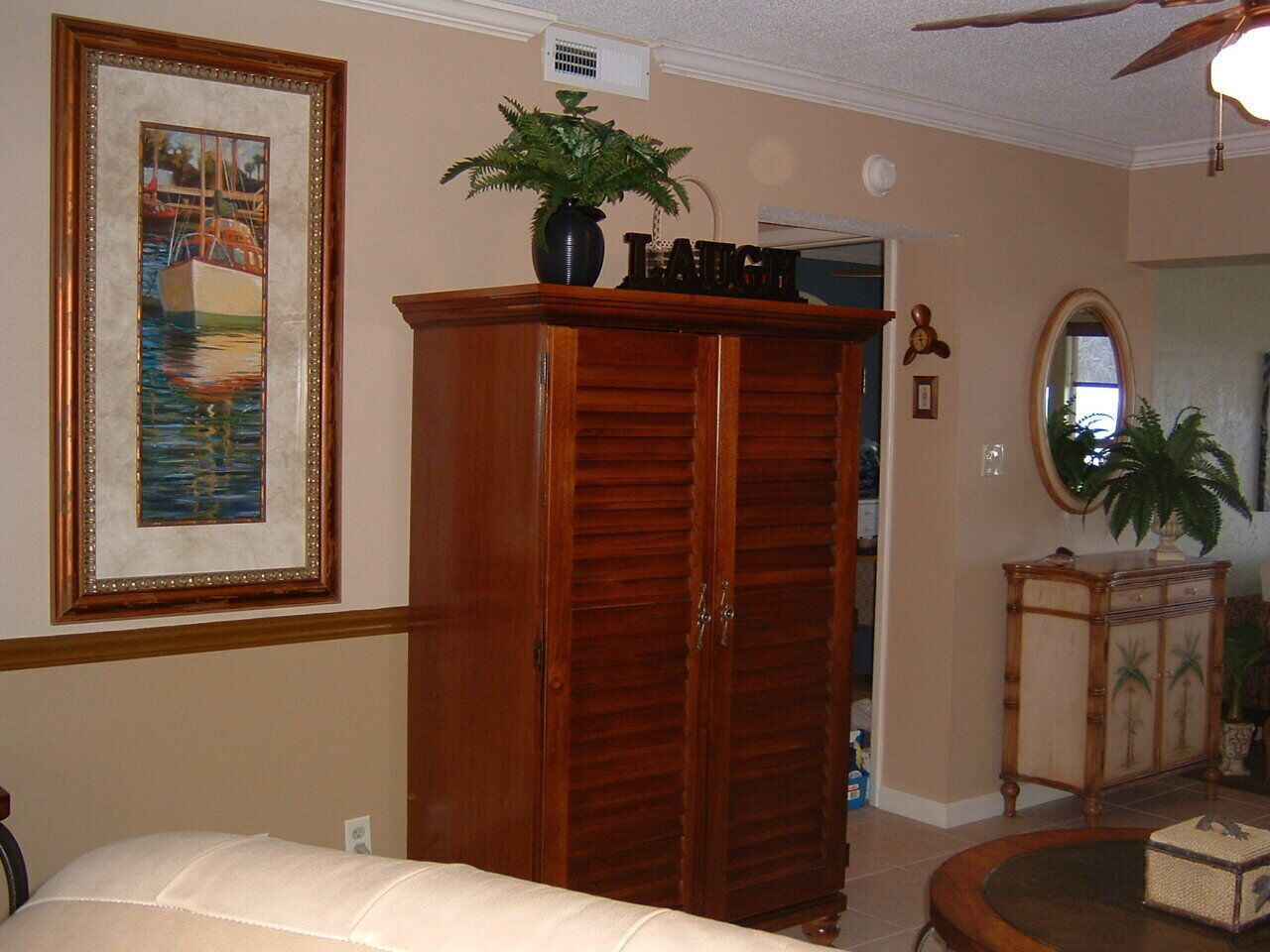 A room with a wooden wardrobe, a framed painting, a potted plant, a mirror, and a small table.