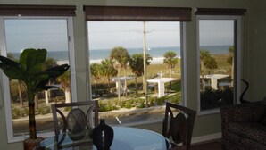 A dining area with a glass table, chairs, a potted plant, and a view of the beach and ocean through large windows. - Stunning Sunset Views-Steps to Siesta Key Beach-A Couples' Romantic Beach Condo (Siesta Key)