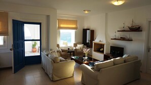 Ein Wohnzimmer mit einer Couch, Sesseln, einem Couchtisch, einem Fernseher und einem Kamin. Ein Fenster bietet Blick ins Freie.