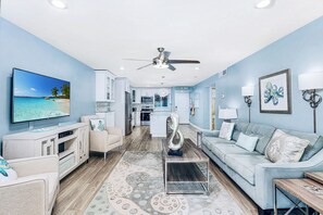 A modern living room with a flat-screen TV, a ceiling fan, a sofa, a coffee table, and a kitchen in the background.